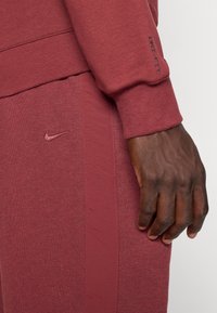 Bordeaux Nike sweatpants med struktureret stof, synlige sømme og et subtilt logo. Indeholder et nærbillede af manchet og hånddetaljer.