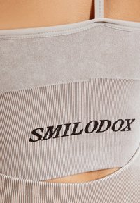 Top corto de canalé gris claro con detalles recortados, con el logotipo "SMILODOX" en negro cosido en la parte frontal. Tejido elástico.