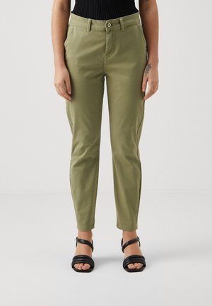 Person iført olivengrønne bukser med lige ben og sorte sandaler med åben tå, stående mod en ensfarvet lys baggrund.