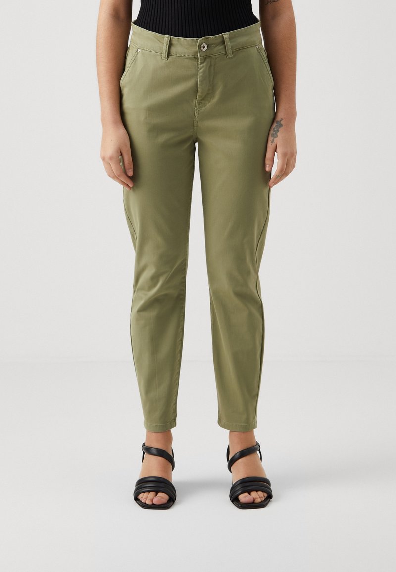 Personne portant un pantalon droit vert olive et des sandales noires à bouts ouverts, debout devant un fond uni clair.
