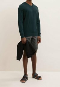Dunkelgrüner V-Ausschnitt-Pullover, graue Jeansshorts, schwarze Kapuzenjacke und dunkle Sandalen. Glatte Textur, lockere Passform und lässiger Stil.