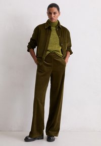 Set od khaki korduroj košulje i hlača; rebrasta tekstura, visoki ovratnik, opušteni kroj. Crne cipele upotpunjuju odjevnu kombinaciju na neutralnoj pozadini.