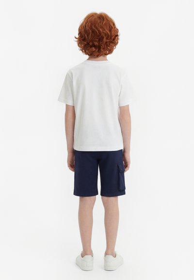 Enfant aux cheveux roux bouclés portant une chemise blanche à manches courtes, un short cargo bleu marine et des baskets blanches, debout de dos sur un fond blanc.