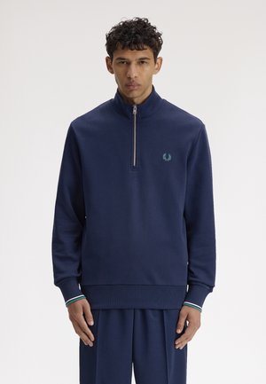 Jeune homme portant un sweat-shirt bleu marine à fermeture éclair partielle avec un petit logo vert sur la poitrine et un pantalon bleu marine assorti, debout devant un fond blanc.