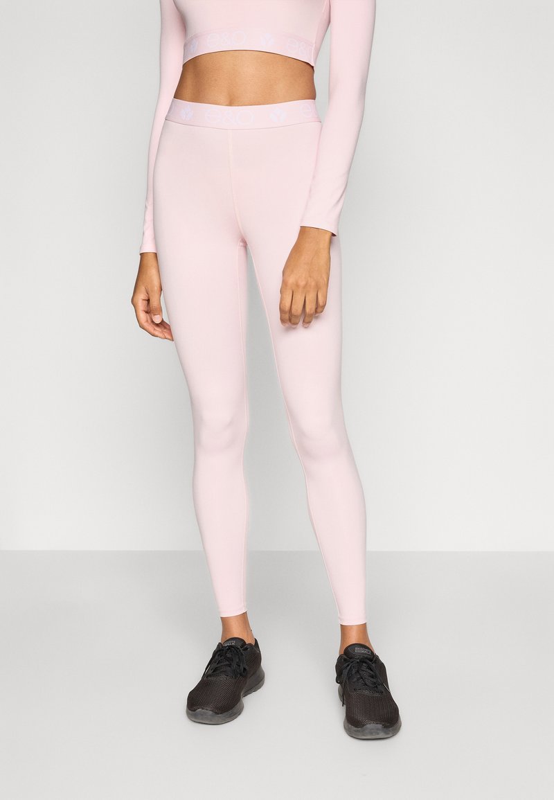 Leggings de sport roses en tissu extensible, avec une taille haute et un design sans coutures. Portés avec des chaussures de sport noires.