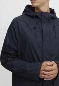 Marine vanntett jakke med hette, med frontlukking, justerbare snor, og en glatt, teksturert overflate.