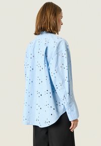 Lichtblauw overhemd met knoopsluiting en uitgesneden details, subtiele textuur; voorzien van lange mouwen en een afgeronde zoom. Draagt bij donkere broek.