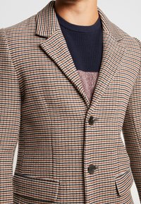 Blazer de lana con un patrón de jaquard en marrón, azul marino y blanco, con solapas de muesca, dos botones y bolsillos laterales.