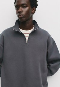 Grauer Zip-Hoodie mit hohem Kragen, lässiger Passform und Frontreißverschluss. Über einem gestreiften weißen Shirt getragen, das aus weichem, strukturiertem Stoff besteht.