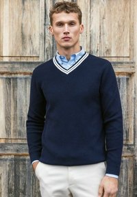 Mörkblå stickad tröja med V-ringning, med vita ränder på kragen, tillsammans med en ljusblå skjorta med krage och krämfärgade byxor.