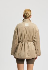 Chaqueta acolchada beige con cuello alto, cintura elástica y tejido texturizado. Presenta una etiqueta rectangular en la parte posterior con la marca.