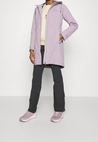Purple waterproof coat with a zip front, hood, and side pockets. Paired with black trousers and light purple sporty shoes.