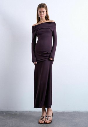 Kvinde står op ad en ensfarvet væg iført en lang, mørk skulderfri kjole med lange ærmer og høje hæle, kigger fremad.