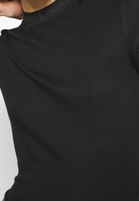 Czarny prążkowany top z długim rękawem i wysokim kołnierzem, z subtelnym logo Calvin Klein na kołnierzu. Gładka tekstura, dopasowany krój.