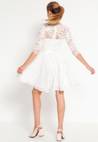 Robe blanche avec un corsage en dentelle, des manches trois-quarts et une jupe plissée. Comprend un détail en forme de clé au dos et est associée à des chaussures à talons bloc beiges.