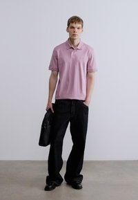 Svetlo roza polo majica, sproščen kroj, kratki rokavi, kombinirana s črnimi širokimi kavbojkami. Model drži črno usnjeno aktovko. Minimalistična ozadje.