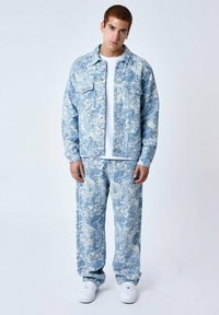Giacca di denim blu con motivi astratti bianchi, abbinata a pantaloni coordinati. Il modello è in piedi su uno sfondo bianco, indossando una T-shirt bianca e sneakers.