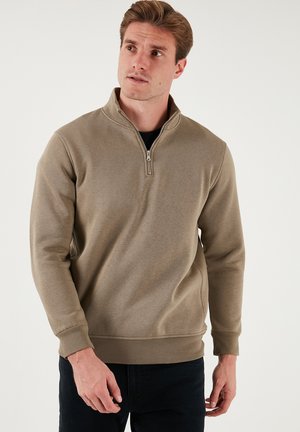 Jasnobrązowa bluza z półzamkiem, z wysokim kołnierzem, wykonana z miękkiego materiału, z ribbowymi mankietami i dołem. Gładka tekstura, minimalistyczny design.