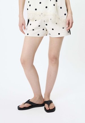 Kvinde iført cremefarvede shorts og top med sorte prikker, sorte flip-flops, stående mod en hvid baggrund.