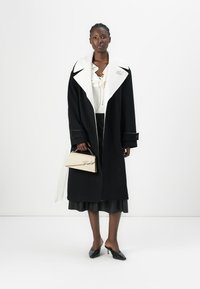 Un long manteau noir en laine avec un grand col blanc, associé à une blouse crème et une jupe noire, tenant un sac à main beige avec un logo.