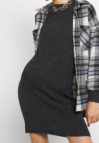Šedé pletené šaty s volným střihem, doplněné modro-bílou kostkovanou flanelovou košilí. Zlatý řetízkový náhrdelník dodává kontrast.