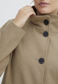 Kameelwollen blend jas met een hoge kraag, voorzien van drie zwarte knopen en een minimalistisch ontwerp. Zachte textuur en strakke lijnen zijn zichtbaar.