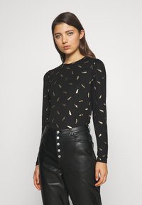 Top preto de mangas longas com um padrão de folhas em dourado. O tecido parece suave, com ligeiro brilho. Combinado com calças de couro pretas de cintura alta.