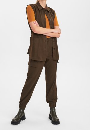 Person trägt eine braune ärmellose, knöpfbare Weste und Cargohosen, ein oranges kurzärmeliges Oberteil und dunkelgrüne klobige Stiefel, steht mit verschränkten Armen.