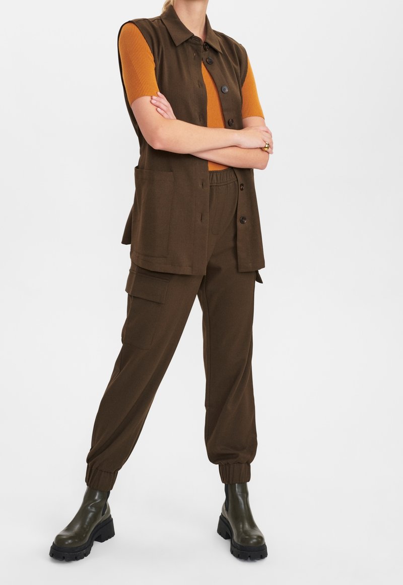 Personne portant un gilet marron sans manches à boutons et un pantalon cargo, un haut orange à manches courtes et des bottes épaisses vert foncé, debout les bras croisés.