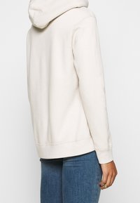 Beiger Kapuzenpullover mit weicher Textur, ausgestattet mit einer Kängurutasche und gerippten Bündchen. Dazu kombiniert sind Jeans aus Denim.