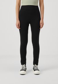 Leggings noirs au design ajusté, fabriqués en tissu lisse. Taille haute et coupe longueur cheville. Associés à des baskets blanches et noires.