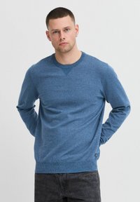 Blauwe gebreide trui met een ronde hals, lange mouwen en een textuurpatroon. Heeft een klein logo aan de onderkant aan de linkerkant.