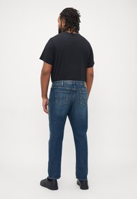 Man with dreadlocks standing facing away wearing black t-shirt, blue jeans, and black sneakers against white background.