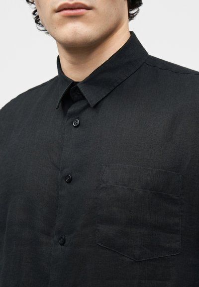 Άνδρας που φοράει μαύρο πουκάμισο με κουμπιά, με μυτερό γιακά και τσέπη στο στήθος, εμφανιζόμενος από τον λαιμό μέχρι το μέσον του κορμού.