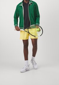 Vihreä vetoketjullinen urheilu takki, musta t-paitaa, keltaiset shortsit ja valkoiset lenkkarit. Pitelee tennismaila ja palloa.