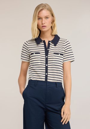 Femme blonde portant une chemise boutonnée à manches courtes rayée bleu marine et blanc avec deux poches avant, rentrée dans un pantalon taille haute bleu marine.