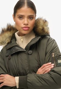 Olijfgroene parka met een met bont gevoerde capuchon, ritssluiting en knoopsluitingen, geribbelde kraag en zijzakken. Bevat een gestructureerde stof en een logo patch.