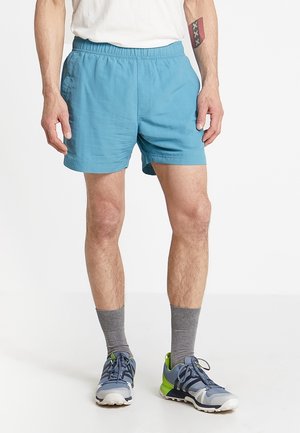 Homme portant un short de sport bleu clair, des chaussettes grises mi-mollet et des chaussures de course grises et vertes, debout les mains détendues le long du corps.