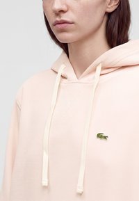 Femme portant un sweat à capuche rose clair avec des cordons blancs et un petit logo crocodile vert sur la poitrine.