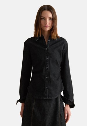 Femme portant une chemise cintrée en jean noir à boutons et une jupe assortie, debout devant un fond clair uni.