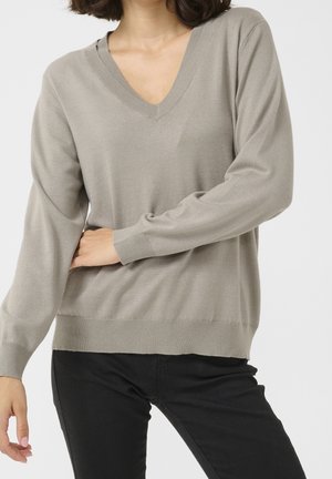 Vrouw draagt een lichtgrijze V-hals trui met lange mouwen en zwarte broek, poseert met één arm over haar taille tegen een effen achtergrond.