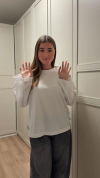 Bluza albă cu mâneci lungi, decolteu ondulat și mâneci largi, asortată cu pantaloni gri închis. Fundalul prezintă dulapuri de culoare deschisă.