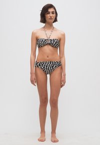 Γυναίκα όρθια, ξυπόλητη, φοράει μπικίνι μπαντό με ασπρόμαυρο πουά και λουρί λαιμού, σε απλό φωτεινό φόντο.