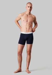 Chiloți boxer de culoare bleu marin, cu un brâu alb cu logo-ul Calvin Klein, design ajustat și material neted; modelul este fără cămașă, stând cu mâinile pe şolduri.