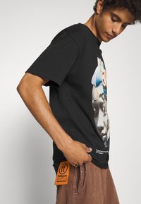 Sort bomulds t-shirt med et levende grafisk print på fronten. Etiketten "240gsm" er synlig hængende fra brune bukser.