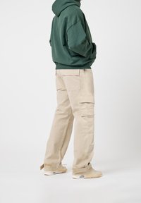 Pantalon cargo beige avec plusieurs poches, associé à un sweat à capuche vert. Les deux vêtements ont une coupe décontractée et sont fabriqués en tissu doux et respirant.