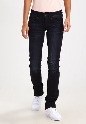 Jeans de mezclilla oscuros de corte ajustado, con un diseño clásico de cinco bolsillos y un ligero desgaste. Combinados con zapatillas blancas.