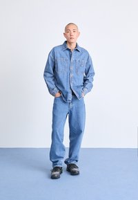 Denimskjorta och byxor i ljusblått, med två bröstfickor, knäppning fram och avslappnad passform, tillsammans med svarta sneakers.