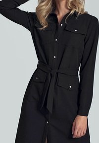 Schwarzes Langarmhemdkleid mit zwei Brusttaschen, Knopfverschluss vorne und einem Kordelzug in der Taille, das eine glatte Textur und ein minimalistisches Design aufweist.