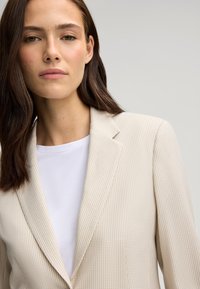 Beżowy żakiet w wzór houndstooth o dopasowanym kroju, z wyciętymi klapami i gładką białą koszulą pod spodem. Naturalne światło podkreśla szczegóły.